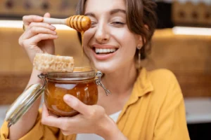 Woman enjoying raw honey, holding a jar with honeycomb and a wooden dipper, highlighting natural honey for women’s wellness and health.