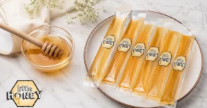 Here is the optimized English Alt Text for "Honey Straws.webp" within your 90-character limit: Six honey sticks on a plate next to a small glass bowl of honey with a dipper.