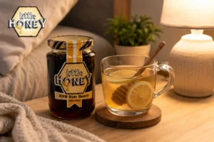 A glass mug of warm honey lemon tea with a wooden dipper inside, sitting next to a jar of Little Honey Sidr Honey on a cozy wooden table.
